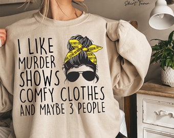 May include: Beige sweatshirt with a graphic of a woman with a messy bun wearing sunglasses and a yellow crime scene tape headband. The text on the sweatshirt reads "I like murder shows comfy clothes and maybe 3 people".