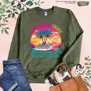 Puede incluir: Sudadera verde militar con un gr&aacute;fico colorido. El gr&aacute;fico presenta un crucero, palmeras y el texto "ANNIVERSARY CRUISE 2026" y "I LOVE IT WHEN WE'RE CRUISIN TOGETHER". La sudadera se combina con vaqueros azules y un bolso tote marr&oacute;n.