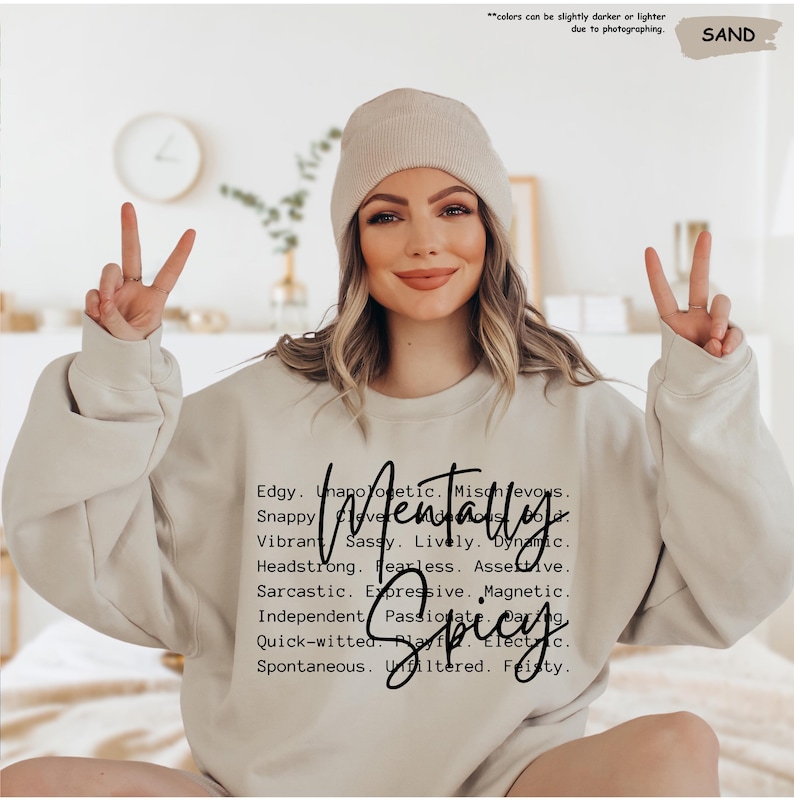 May include: A sand-colored sweatshirt with the words "Mentally Spicy" in a handwritten font. The sweatshirt also lists several adjectives. The model is wearing a matching beanie and making a peace sign with both hands.