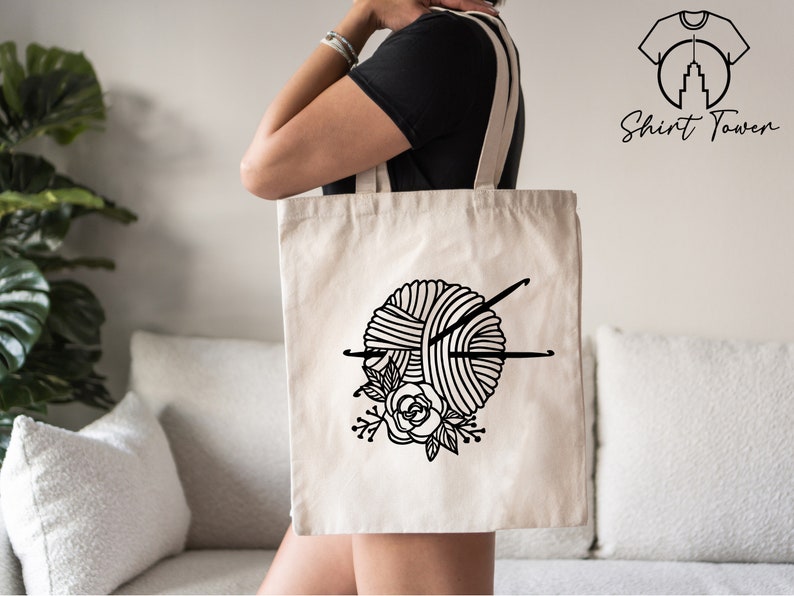 Pu&ograve; includere: Una borsa tote in tela bianca con un'illustrazione in bianco e nero di un gomitolo di lana e ferri da maglia con accenti floreali. La borsa ha manici neri.