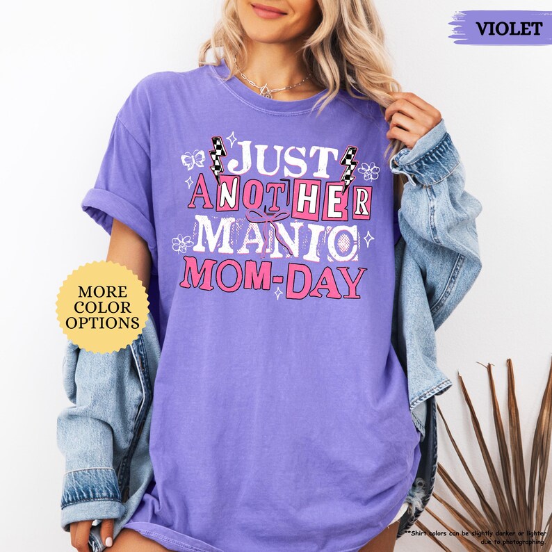 May include: A violet-colored t-shirt with the text "JUST ANOTHER MANIC MOM-DAY" in a pink and white design. The shirt has short sleeves and a relaxed fit. The words "MORE COLOR OPTIONS" are also visible.