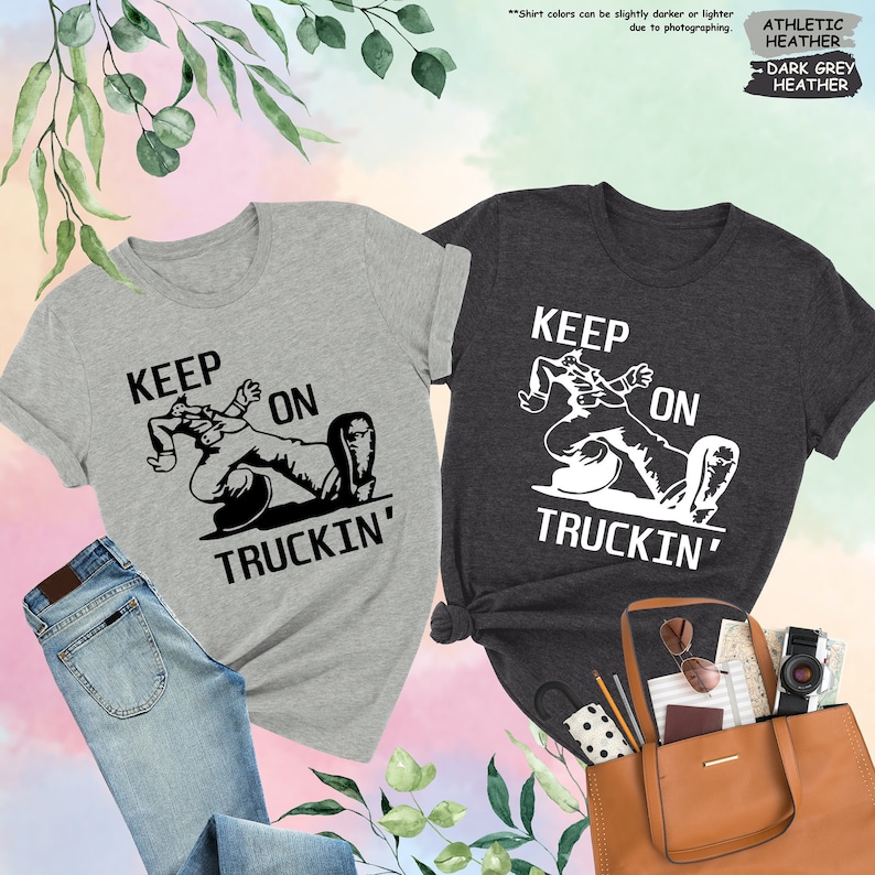 May include: Two t-shirts, one gray and one dark gray, each with the phrase "KEEP ON TRUCKIN'" in white block letters and a graphic of a person reclining on a truck tire. The gray shirt is paired with blue jeans. The dark gray shirt is next to a brown tote bag.