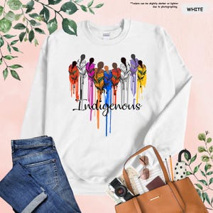 May include: White sweatshirt with a colorful design of stylized figures with feathers and the word "Indigenous" in a dripping paint style.