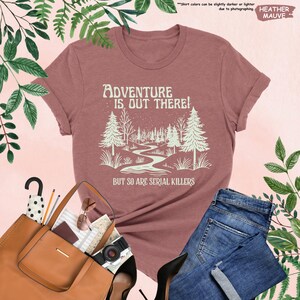Op de afbeelding: Mauvekleurig T-shirt met de tekst "ADVENTURE IS OUT THERE! BUT SO ARE SERIAL KILLERS" en een bosafbeelding. Het shirt wordt gecombineerd met een blauwe spijkerbroek, een bruine handtas en zwarte hakken.