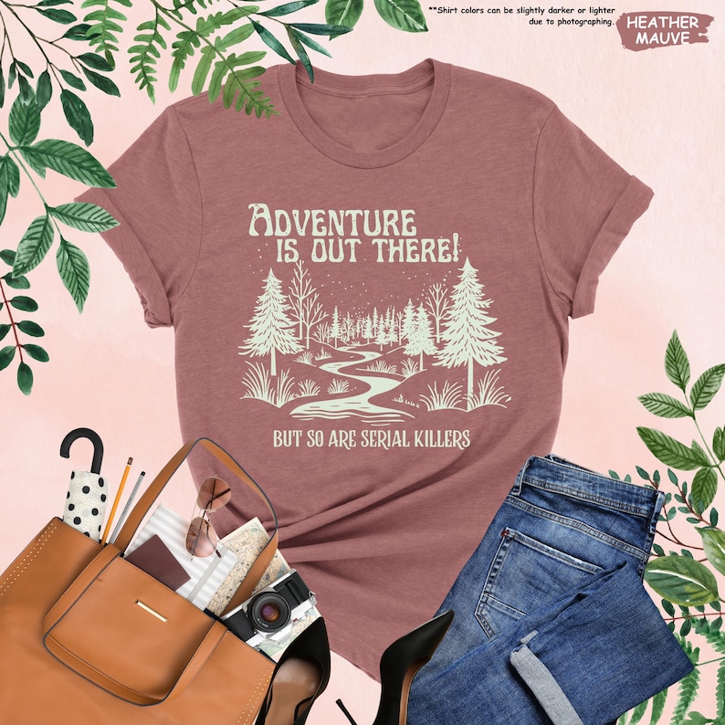 Op de afbeelding: Mauvekleurig T-shirt met de tekst "ADVENTURE IS OUT THERE! BUT SO ARE SERIAL KILLERS" en een bosafbeelding. Het shirt wordt gecombineerd met een blauwe spijkerbroek, een bruine handtas en zwarte hakken.