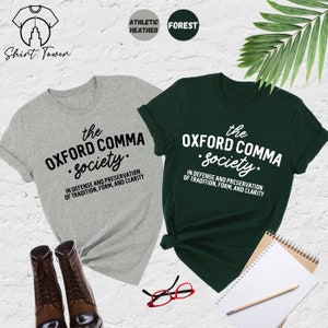 Op de afbeelding: Twee T-shirts met de tekst "the Oxford comma society." in zwart en wit. De tekst staat op een grijs en een groen T-shirt. De tekst luidt "In verdediging en behoud van traditie, vorm en duidelijkheid."