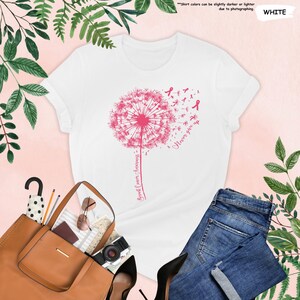 Puede incluir: Camiseta blanca con un dise&ntilde;o de diente de le&oacute;n rosa y las palabras "Breast Cancer Awareness" y "Never give up." La camiseta se combina con vaqueros azules, un bolso tote marr&oacute;n y tacones negros. El dise&ntilde;o incluye lazos rosas.