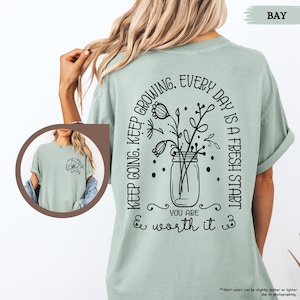 May include: A light green t-shirt with a black graphic design featuring a mason jar with flowers and the text "Keep going. Keep growing. Every day is a fresh start. You are worth it."