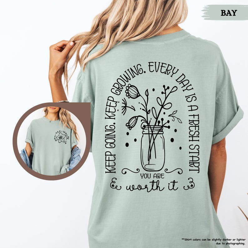 May include: A light green t-shirt with a black graphic design featuring a mason jar with flowers and the text "Keep going. Keep growing. Every day is a fresh start. You are worth it."
