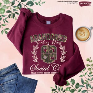 May include: Maroon sweatshirt with the text "ABANDONED Hunting Wives Social Club" and hunting-themed graphics. The sweatshirt is folded with a pair of blue jeans and a cup of coffee. The text "HELLO HUNTING SEASON. GOOD..." is also visible.