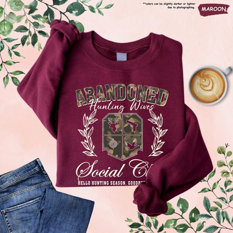 May include: Maroon sweatshirt with the text "ABANDONED Hunting Wives Social Club" and hunting-themed graphics. The sweatshirt is folded with a pair of blue jeans and a cup of coffee. The text "HELLO HUNTING SEASON. GOOD..." is also visible.