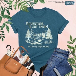 Op de afbeelding: Heather deep teal t-shirt met de witte tekst "Adventure is Out There! But So Are Serial Killers". De afbeelding toont een boslandschap met een kronkelende rivier. Opgerolde mouwen en een ronde hals.