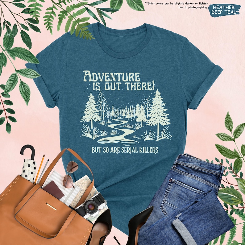 Op de afbeelding: Heather deep teal t-shirt met de witte tekst "Adventure is Out There! But So Are Serial Killers". De afbeelding toont een boslandschap met een kronkelende rivier. Opgerolde mouwen en een ronde hals.