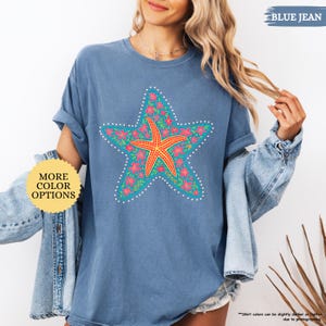 May include: Blue jean colored t-shirt featuring a large starfish design. The starfish is orange, surrounded by a turquoise star shape with pink flowers and white dots. The shirt has the text "MORE COLOR OPTIONS" and "BLUE JEAN".