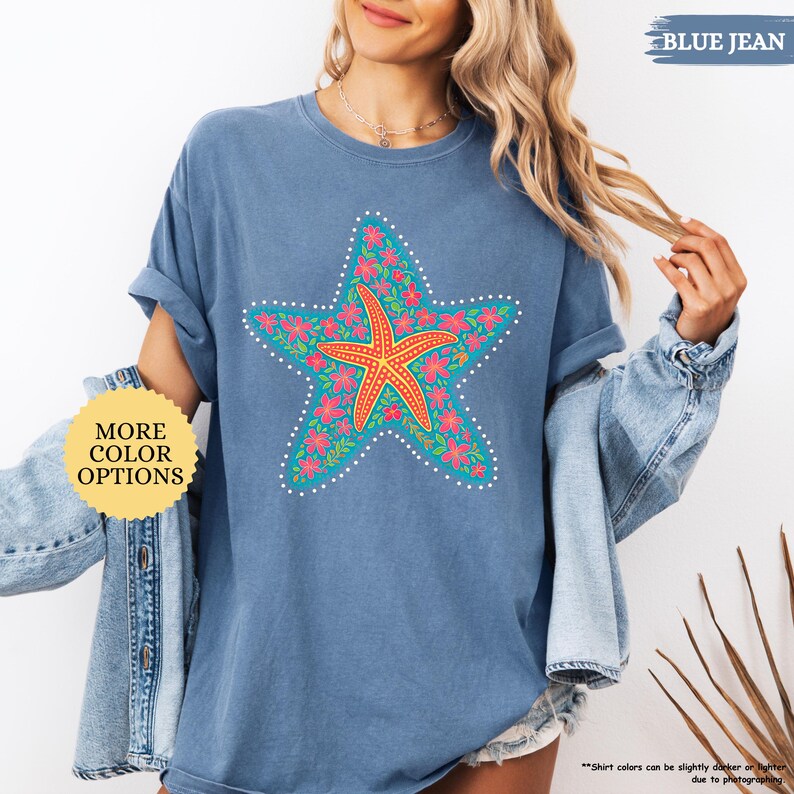 May include: Blue jean colored t-shirt featuring a large starfish design. The starfish is orange, surrounded by a turquoise star shape with pink flowers and white dots. The shirt has the text "MORE COLOR OPTIONS" and "BLUE JEAN".