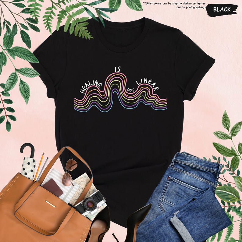 Op de afbeelding: Zwart T-shirt met de tekst "HEALING IS NOT LINEAR" in een kleurrijk, golvend ontwerp. Het shirt wordt gecombineerd met een blauwe jeans, een bruine totebag en accessoires. De kleur van het shirt is zwart.