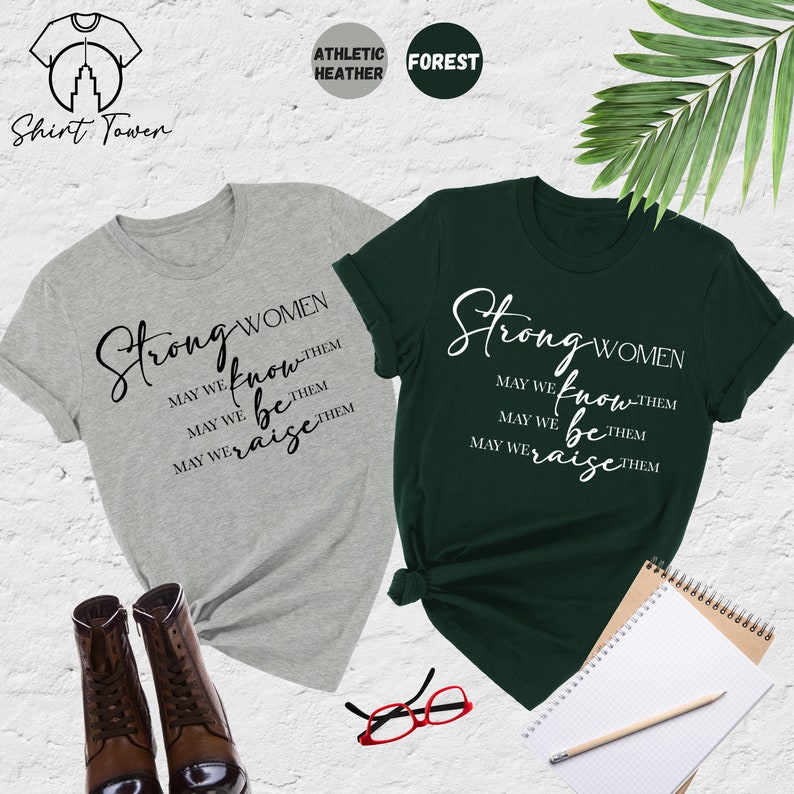 Puede incluir: Dos camisetas con el texto "Strong Women" y la frase "May we know them, May we be them, May we raise them" impresa en ellas. Las camisetas son de color gris jaspeado deportivo y verde bosque.