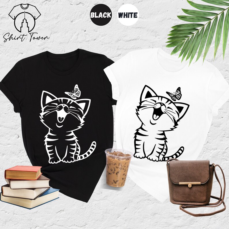 May include: Two t-shirts with a black and white graphic of a smiling cat with a butterfly above it. The t-shirts are available in black and white.