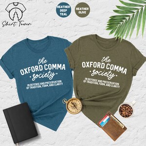 Op de afbeelding: Twee T-shirts met de tekst "the Oxford comma society." in witte letters op een teal en olijfgroene achtergrond. De tekst eronder luidt "In defense and preservation of tradition, form, and clarity."