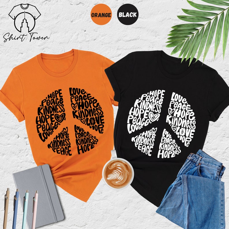 May include: Two t-shirts with a peace sign design. The peace sign is made up of the words "Hope", "Peace", "Love", and "Kindness". One shirt is orange and the other is black.