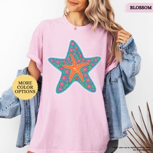 May include: A light pink t-shirt featuring a large starfish design. The starfish is teal with a floral pattern and a central orange starfish. The shirt has the text "BLOSSOM" and "MORE COLOR OPTIONS".