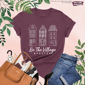 May include: A heather maroon t-shirt with a white graphic of three houses and the text "Be The Village #FOSTERO".