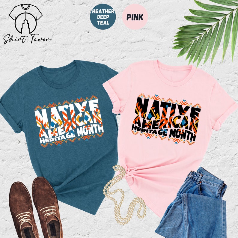 May include: Two t-shirts, one in heather deep teal and one in pink, with the text 'Native American Heritage Month' in colorful block letters with geometric patterns.