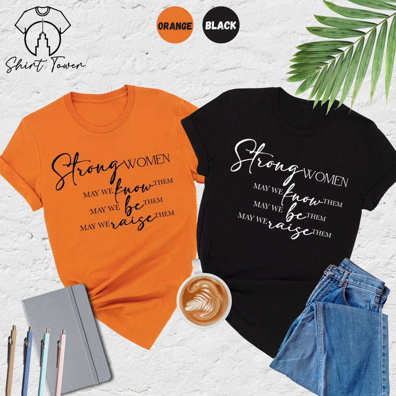 Puede incluir: Dos camisetas, una naranja y otra negra, con el texto "Strong Women" y la frase "May we know them, May we be them, May we raise them".
