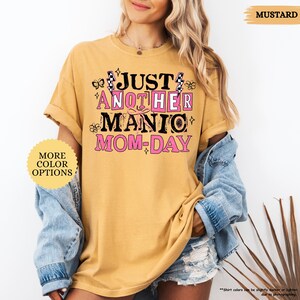 May include: Mustard-colored t-shirt with the text "JUST ANOTHER MANIC MOM-DAY" in black and pink. The shirt has rolled-up sleeves and is paired with a denim jacket. The words "MORE COLOR OPTIONS" are also visible.