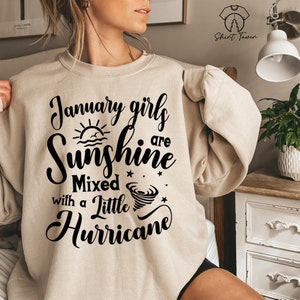 May include: A beige sweatshirt with black text that reads "January girls are sunshine mixed with a little hurricane". The text includes a sun, waves, stars, and a tornado graphic.