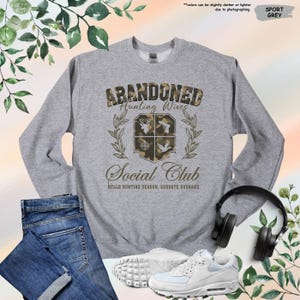 May include: Sport grey sweatshirt with the text "ABANDONED Hunting Wives Social Club". The design includes a crest with hunting-themed graphics. The phrase "Hello Hunting Season, Goodbye Husband" is printed below the club name. A pair of jeans, sneakers, and headphones are also in the image.