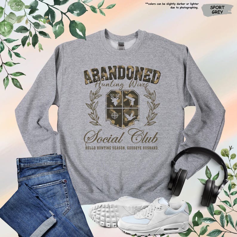 May include: Sport grey sweatshirt with the text "ABANDONED Hunting Wives Social Club". The design includes a crest with hunting-themed graphics. The phrase "Hello Hunting Season, Goodbye Husband" is printed below the club name. A pair of jeans, sneakers, and headphones are also in the image.