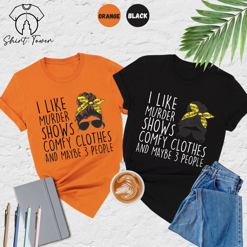 May include: Two t-shirts with a graphic of a woman with a messy bun and sunglasses. The text on the shirts reads "I like murder shows, comfy clothes and maybe 3 people". The shirts are available in orange and black.