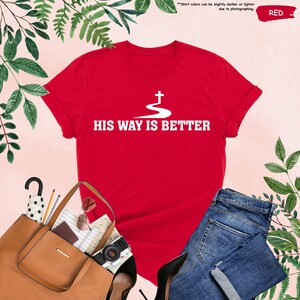 May include: A red t-shirt with the text "HIS WAY IS BETTER" in white, along with a white cross design. The shirt is displayed with a pair of blue jeans, a brown purse, and other accessories.