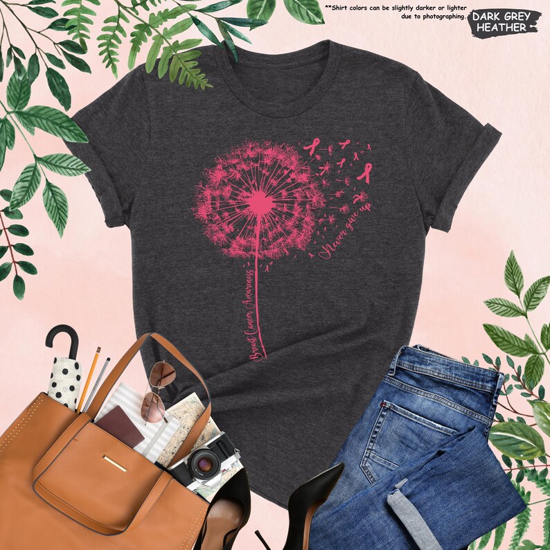 Puede incluir: Camiseta gris oscuro jaspeado con un dise&ntilde;o de diente de le&oacute;n rosa y las palabras "Breast Cancer Awareness" y "Never give up". La imagen incluye vaqueros azules, un bolso tote marr&oacute;n y tacones negros.