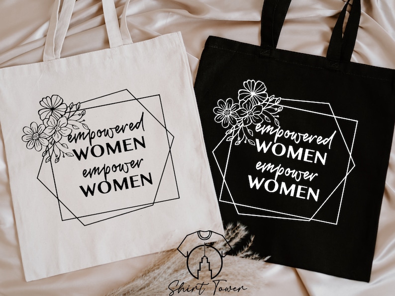 May include: Two tote bags featuring the text "empowered women empower women" within a black geometric frame and a floral design. One bag is white with black text and the other is black with white text.