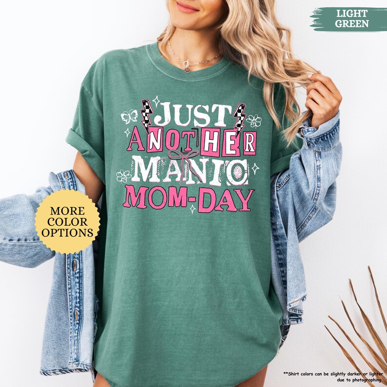 May include: A light green t-shirt with the text "JUST ANOTHER MANIC MOM-DAY" in white and pink. The shirt has short sleeves and a crew neck. A yellow circle with the words "MORE COLOR OPTIONS" is on the left.