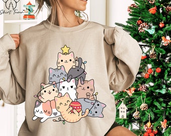 May include: Beige sweatshirt with a Christmas tree design made of cartoon cats. The cats are in various colours, with a star on top and string lights. The sweatshirt has the Shirt Tower logo in the upper left corner.