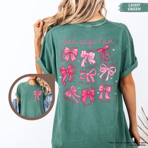K&ouml;nnte beinhalten: Ein hellgr&uuml;nes T-Shirt mit dem Text "God says I am" auf der R&uuml;ckseite. Der Text ist von rosa Schleifen mit Bibelversen umgeben.