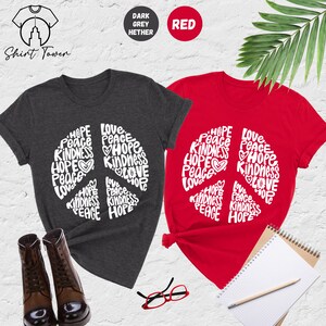 May include: Two t-shirts with a peace sign design. The peace sign is made up of the words "Hope", "Peace", "Love", and "Kindness". One shirt is dark heather grey and the other is red.