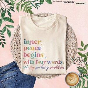 May include: A beige t-shirt with the text "inner peace begins with four words: not my fucking problem" printed in colourful letters. The text is surrounded by small flower graphics.