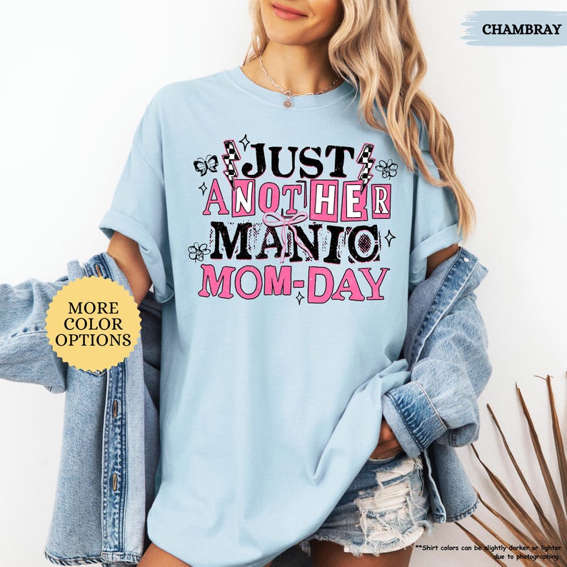 May include: Light blue t-shirt with the text "JUST ANOTHER MANIC MOM-DAY" in black and pink. The design includes butterflies, flowers, and lightning bolts. The shirt is a chambray color and has "MORE COLOR OPTIONS" text on a yellow circle.