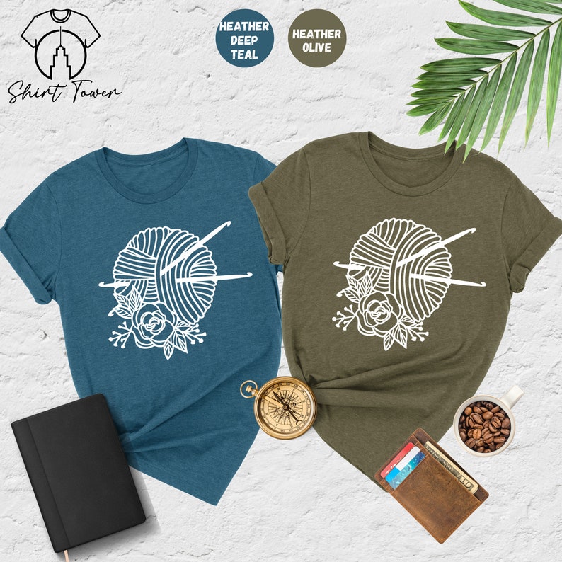 Pode incluir: Duas camisetas com um gr&aacute;fico branco de um novelo de l&atilde; com agulhas de tric&ocirc; e um design floral. A camiseta &agrave; esquerda &eacute; azul-petr&oacute;leo heather e a camiseta &agrave; direita &eacute; oliva heather.