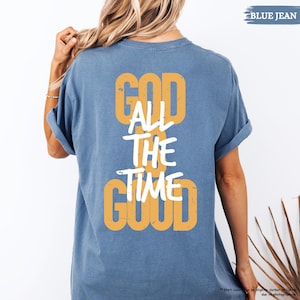 May include: Blue jean-colored t-shirt with the text "GOD ALL THE TIME GOOD" in a combination of yellow and white. The shirt has short sleeves and a relaxed fit. The words are stacked vertically on the back of the shirt.