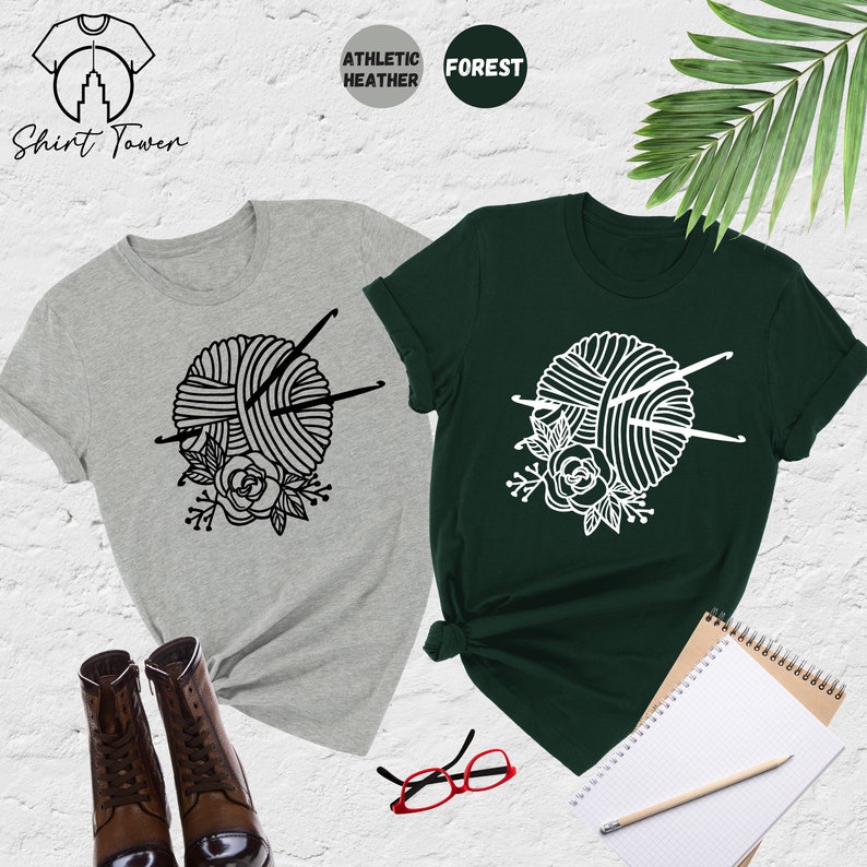 Pode incluir: Duas camisetas com um gr&aacute;fico de um novelo de l&atilde; com agulhas de tric&ocirc; e flores. A camiseta &agrave; esquerda &eacute; cinza e a camiseta &agrave; direita &eacute; verde. "Shirt Town" est&aacute; escrito no canto superior esquerdo da imagem.