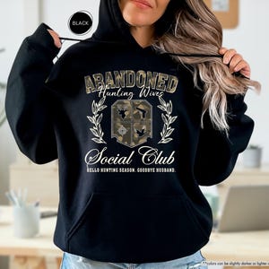 May include: Black hooded sweatshirt with the text "ABANDONED Hunting Wives Social Club" in a camouflage design. The design includes a shield with hunting-related icons and the phrase "HELLO HUNTING SEASON, GOODBYE HUSBAND."