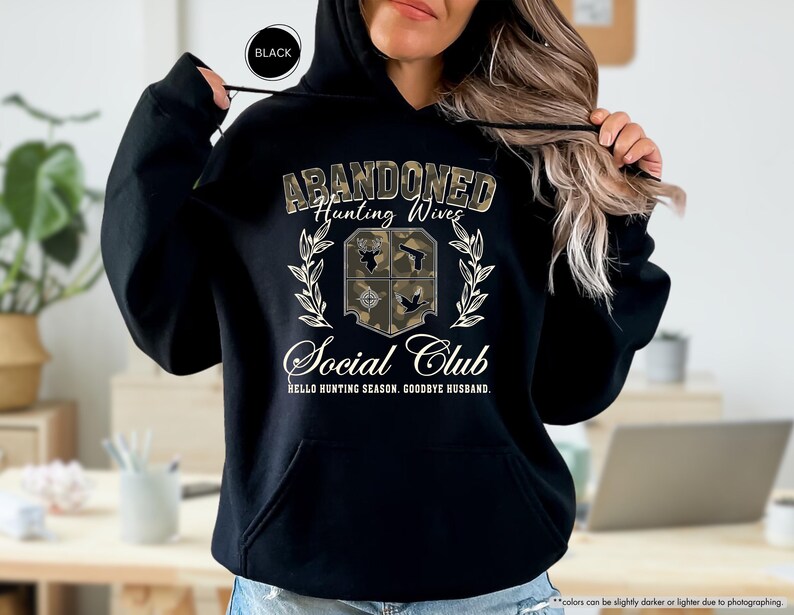 May include: Black hooded sweatshirt with the text "ABANDONED Hunting Wives Social Club" in a camouflage design. The design includes a shield with hunting-related icons and the phrase "HELLO HUNTING SEASON, GOODBYE HUSBAND."