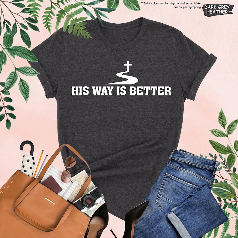May include: Dark grey heather t-shirt with the text "HIS WAY IS BETTER" in white. The shirt features a graphic of a cross above a winding path. The shirt is paired with blue jeans, a brown purse, and black heels.