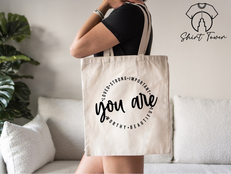 K&ouml;nnte beinhalten: Eine wei&szlig;e Canvas-Tragetasche mit schwarzem Text, der "You are loved, strong, important, worthy, beautiful" lautet.