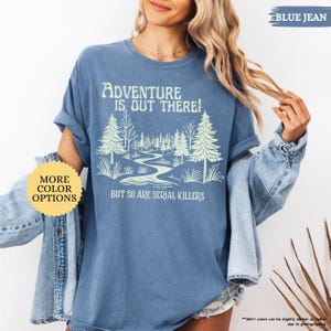 May include: Blue jean-colored t-shirt with the text "Adventure is Out There!" above a graphic of a winding river and trees. Below the graphic, the text reads "But So Are Serial Killers." A yellow circle with "More Color Options" is on the left.